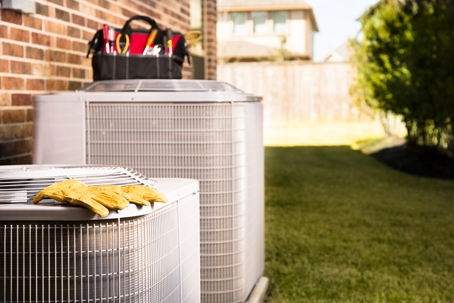 Bag of repairman's work tools, gloves on top of air conditioner units outside a brick home.